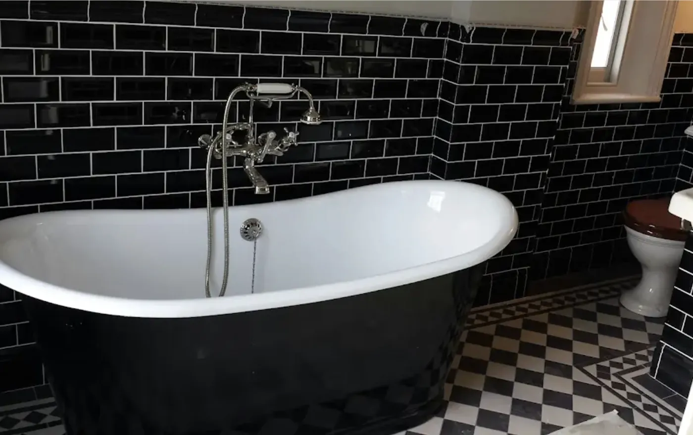 Bathroom tiled in black metro brick with a black roll-top bath and black and white checkerboard floor
